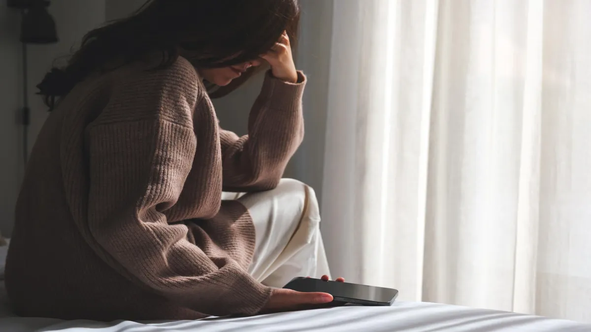 Frau sitzt nachdenklich am Bett, Smartphone in der Hand, Licht durch Vorhang