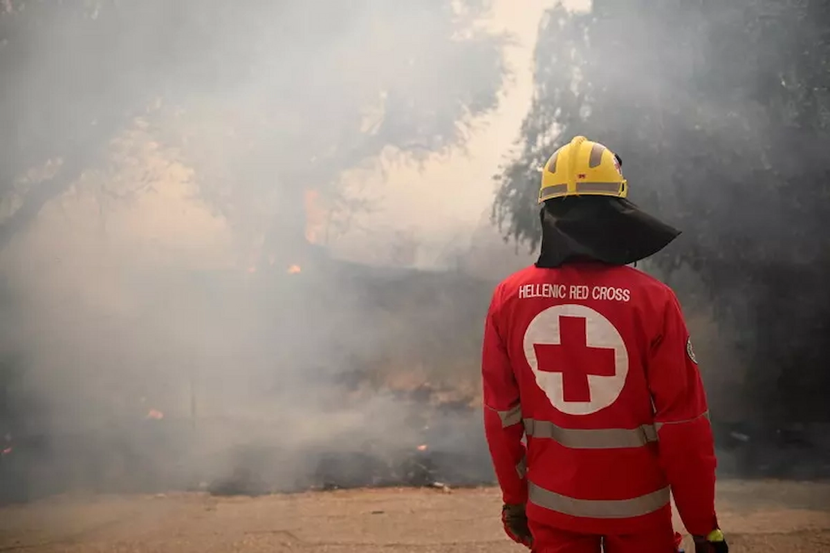 Mitglied des Hellenischen Roten Kreuzes in Schutzkleidung blickt auf Rauch und Flammen bei einem Waldbrand in Griechenland.
